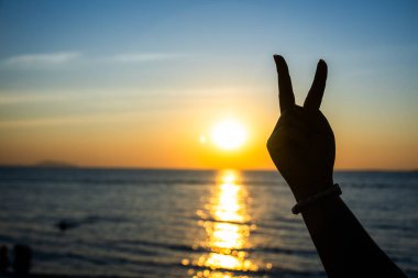 Siluetli el, okyanusun üzerinde parlayan bir günbatımına karşı barış işareti yapıyor. Altın ışık suya yansıyor.