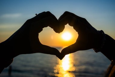 Siluetli eller, okyanusun üzerinde altın bir günbatımına karşı bir kalp şekli oluşturuyor. Güneş ışığı, sakin suya yansıyor..