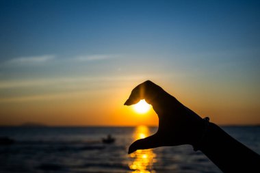 Silüetli el, okyanusun üzerinde parlayan bir günbatımının önünde, altın güneş ışığını yansıtan yarım kalp jesti oluşturuyor.