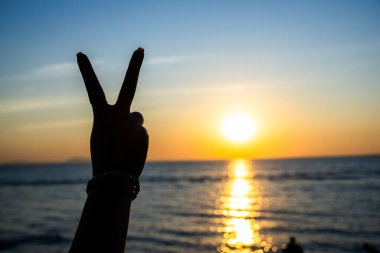 Siluetli el, okyanusun üzerinde parlayan bir günbatımına karşı barış işareti yapıyor. Altın ışık suya yansıyor.