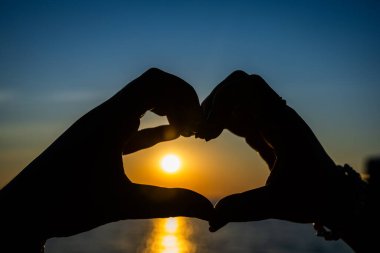 Siluetli eller, okyanusun üzerinde altın bir günbatımına karşı bir kalp şekli oluşturuyor. Güneş ışığı, sakin suya yansıyor..