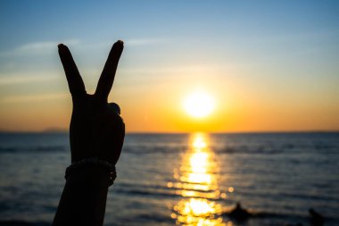 Siluetli el, okyanusun üzerinde parlayan bir günbatımına karşı barış işareti yapıyor. Altın ışık suya yansıyor.