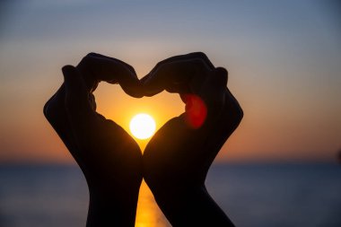 Siluetli eller bir kalp şekli oluşturuyor. Okyanusun üzerinde batarken parlayan güneşi mükemmel bir şekilde çerçeveliyor. Huzurlu bir arkaplan renkleri ve durgun sularla..