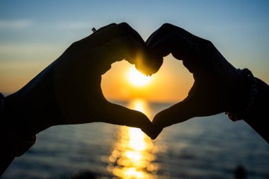 Siluetli eller, okyanusun üzerinde altın bir günbatımına karşı bir kalp şekli oluşturuyor. Güneş ışığı, sakin suya yansıyor..