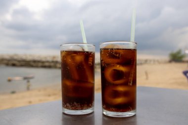Bir bardak buzlu kola, deniz kenarındaki bir masada, bulutlu bir arka planda duruyor. İçkinin kabarcıkları ve yoğunlaşması, canlandırıcı kalitesini vurguluyor. Plaj manzarası rahatlatıcı bir yaz havası yaratıyor..