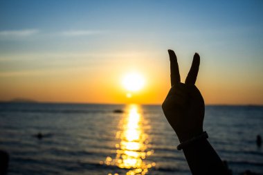 Siluetli el, okyanusun üzerinde parlayan bir günbatımına karşı barış işareti yapıyor. Altın ışık suya yansıyor.
