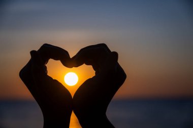 Siluetli eller bir kalp şekli oluşturuyor. Okyanusun üzerinde batarken parlayan güneşi mükemmel bir şekilde çerçeveliyor. Huzurlu bir arkaplan renkleri ve durgun sularla..