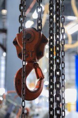 Fabrika ya da atölye ortamında asılı ağır metal zincirleri olan kırmızı sanayi zincirinin yakın çekimi. Bulanık arkaplan makine ve ekipmanları gösterir