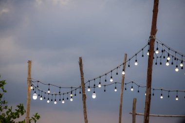 Alacakaranlık gökyüzüne karşı kırsal ahşap direklerin arasında asılı duran dış mekan telli ışıklar. Ampullerin sıcaklığı, alacakaranlık gökyüzünün serin tonlarıyla çelişiyor. Sıcak ve davetkar bir atmosfer yaratıyor..