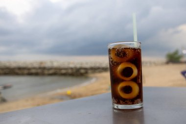 Bir bardak buzlu kola, deniz kenarındaki bir masada, bulutlu bir arka planda duruyor. İçkinin kabarcıkları ve yoğunlaşması, canlandırıcı kalitesini vurguluyor. Plaj manzarası rahatlatıcı bir yaz havası yaratıyor..