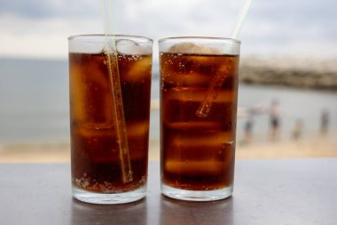 Bir bardak buzlu kola, deniz kenarındaki bir masada, bulutlu bir arka planda duruyor. İçkinin kabarcıkları ve yoğunlaşması, canlandırıcı kalitesini vurguluyor. Plaj manzarası rahatlatıcı bir yaz havası yaratıyor..