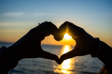 Siluetli eller, okyanusun üzerinde altın bir günbatımına karşı bir kalp şekli oluşturuyor. Güneş ışığı, sakin suya yansıyor..