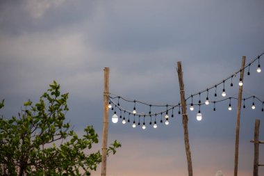 Alacakaranlık gökyüzüne karşı kırsal ahşap direklerin arasında asılı duran dış mekan telli ışıklar. Ampullerin sıcaklığı, alacakaranlık gökyüzünün serin tonlarıyla çelişiyor. Sıcak ve davetkar bir atmosfer yaratıyor..