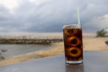 Bir bardak buzlu kola, deniz kenarındaki bir masada, bulutlu bir arka planda duruyor. İçkinin kabarcıkları ve yoğunlaşması, canlandırıcı kalitesini vurguluyor. Plaj manzarası rahatlatıcı bir yaz havası yaratıyor..