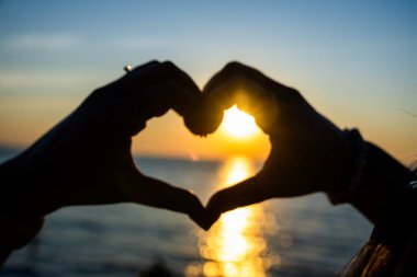 Siluetli eller, okyanusun üzerinde altın bir günbatımına karşı bir kalp şekli oluşturuyor. Güneş ışığı, sakin suya yansıyor..