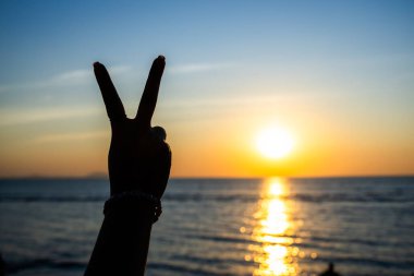 Siluetli el, okyanusun üzerinde parlayan bir günbatımına karşı barış işareti yapıyor. Altın ışık suya yansıyor.