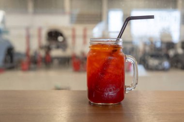 Ahşap bir masaya konmuş siyah pipetle mason kavanozunda servis edilen ferahlatıcı buzlu kırmızı içecek. Arkaplanda bulanık bir görüntü var