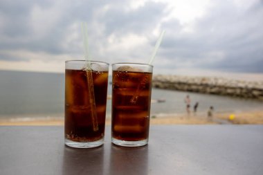 Bir bardak buzlu kola, deniz kenarındaki bir masada, bulutlu bir arka planda duruyor. İçkinin kabarcıkları ve yoğunlaşması, canlandırıcı kalitesini vurguluyor. Plaj manzarası rahatlatıcı bir yaz havası yaratıyor..