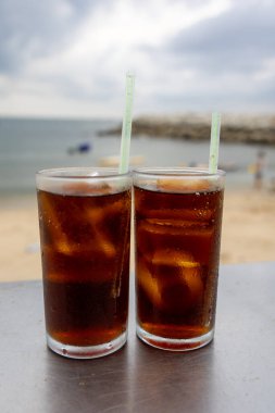 Bir bardak buzlu kola, deniz kenarındaki bir masada, bulutlu bir arka planda duruyor. İçkinin kabarcıkları ve yoğunlaşması, canlandırıcı kalitesini vurguluyor. Plaj manzarası rahatlatıcı bir yaz havası yaratıyor..