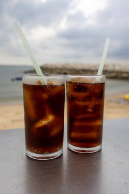 Bir bardak buzlu kola, deniz kenarındaki bir masada, bulutlu bir arka planda duruyor. İçkinin kabarcıkları ve yoğunlaşması, canlandırıcı kalitesini vurguluyor. Plaj manzarası rahatlatıcı bir yaz havası yaratıyor..