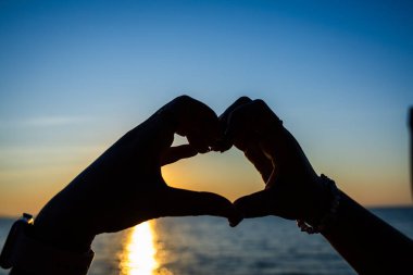 Siluetli eller, okyanusun üzerinde altın bir günbatımına karşı bir kalp şekli oluşturuyor. Güneş ışığı, sakin suya yansıyor..