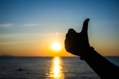Silüetli el, okyanusun üzerinde canlı bir günbatımına karşı baş parmak işareti yapıyor. Altın güneş ışığı suya yansıyor..