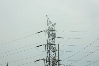 Yüksek voltajlı elektrik hatları taşıyan uzun, çelik kafes şanzıman kulesinin manzarası. Kule gri, bulutlu bir gökyüzüne karşı duruyor..
