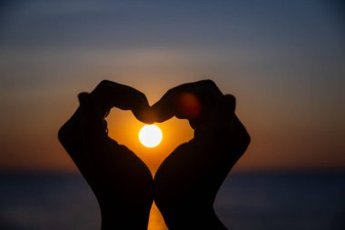 Siluetli eller bir kalp şekli oluşturuyor. Okyanusun üzerinde batarken parlayan güneşi mükemmel bir şekilde çerçeveliyor. Huzurlu bir arkaplan renkleri ve durgun sularla..