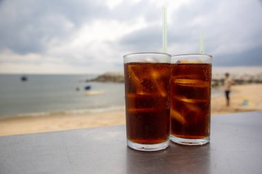 Bir bardak buzlu kola, deniz kenarındaki bir masada, bulutlu bir arka planda duruyor. İçkinin kabarcıkları ve yoğunlaşması, canlandırıcı kalitesini vurguluyor. Plaj manzarası rahatlatıcı bir yaz havası yaratıyor..
