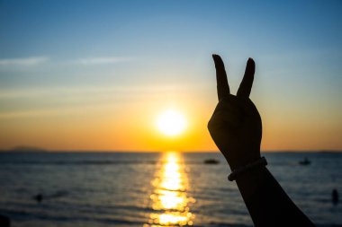 Siluetli el, okyanusun üzerinde parlayan bir günbatımına karşı barış işareti yapıyor. Altın ışık suya yansıyor.