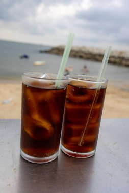 Bir bardak buzlu kola, deniz kenarındaki bir masada, bulutlu bir arka planda duruyor. İçkinin kabarcıkları ve yoğunlaşması, canlandırıcı kalitesini vurguluyor. Plaj manzarası rahatlatıcı bir yaz havası yaratıyor..