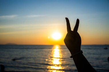 Siluetli el, okyanusun üzerinde parlayan bir günbatımına karşı barış işareti yapıyor. Altın ışık suya yansıyor.