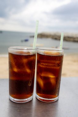 Bir bardak buzlu kola, deniz kenarındaki bir masada, bulutlu bir arka planda duruyor. İçkinin kabarcıkları ve yoğunlaşması, canlandırıcı kalitesini vurguluyor. Plaj manzarası rahatlatıcı bir yaz havası yaratıyor..