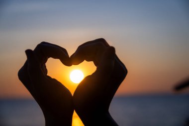 Siluetli eller bir kalp şekli oluşturuyor. Okyanusun üzerinde batarken parlayan güneşi mükemmel bir şekilde çerçeveliyor. Huzurlu bir arkaplan renkleri ve durgun sularla..