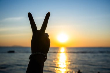 Siluetli el, okyanusun üzerinde parlayan bir günbatımına karşı barış işareti yapıyor. Altın ışık suya yansıyor.