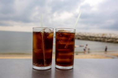 Bir bardak buzlu kola, deniz kenarındaki bir masada, bulutlu bir arka planda duruyor. İçkinin kabarcıkları ve yoğunlaşması, canlandırıcı kalitesini vurguluyor. Plaj manzarası rahatlatıcı bir yaz havası yaratıyor..