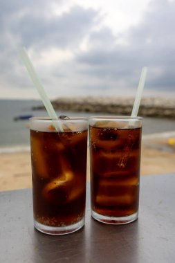 Bir bardak buzlu kola, deniz kenarındaki bir masada, bulutlu bir arka planda duruyor. İçkinin kabarcıkları ve yoğunlaşması, canlandırıcı kalitesini vurguluyor. Plaj manzarası rahatlatıcı bir yaz havası yaratıyor..