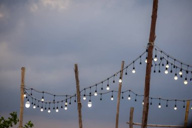 Alacakaranlık gökyüzüne karşı kırsal ahşap direklerin arasında asılı duran dış mekan telli ışıklar. Ampullerin sıcaklığı, alacakaranlık gökyüzünün serin tonlarıyla çelişiyor. Sıcak ve davetkar bir atmosfer yaratıyor..