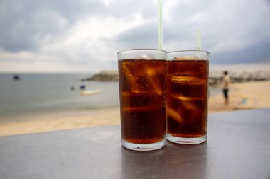 Bir bardak buzlu kola, deniz kenarındaki bir masada, bulutlu bir arka planda duruyor. İçkinin kabarcıkları ve yoğunlaşması, canlandırıcı kalitesini vurguluyor. Plaj manzarası rahatlatıcı bir yaz havası yaratıyor..