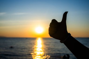 Silüetli el, okyanusun üzerinde canlı bir günbatımına karşı baş parmak işareti yapıyor. Altın güneş ışığı suya yansıyor..