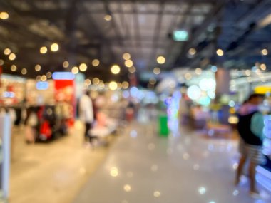 Bir alışveriş merkezinin içini gösteren bulanık bir görüntü. Olay yeri bulanık, belirsiz dükkanlar, ışıklar ve yürüyen insanlarla dolu. Genel izlenim yumuşak ve soyut.