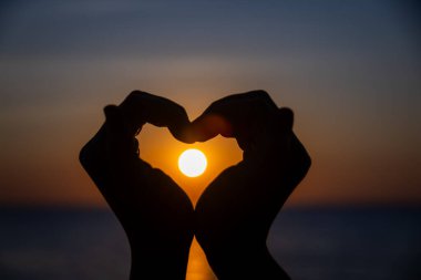 Siluetli eller bir kalp şekli oluşturuyor. Okyanusun üzerinde batarken parlayan güneşi mükemmel bir şekilde çerçeveliyor. Huzurlu bir arkaplan renkleri ve durgun sularla..