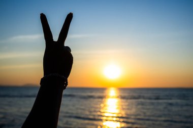 Siluetli el, okyanusun üzerinde parlayan bir günbatımına karşı barış işareti yapıyor. Altın ışık suya yansıyor.