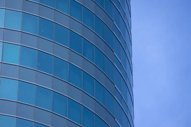 Modern bir binanın birçok dikdörtgen pencereli bir bölümü berrak, parlak mavi gökyüzünü yansıtıyor..