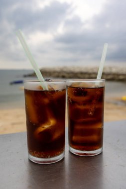 Bir bardak buzlu kola, deniz kenarındaki bir masada, bulutlu bir arka planda duruyor. İçkinin kabarcıkları ve yoğunlaşması, canlandırıcı kalitesini vurguluyor. Plaj manzarası rahatlatıcı bir yaz havası yaratıyor..
