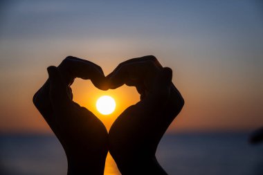 Siluetli eller bir kalp şekli oluşturuyor. Okyanusun üzerinde batarken parlayan güneşi mükemmel bir şekilde çerçeveliyor. Huzurlu bir arkaplan renkleri ve durgun sularla..