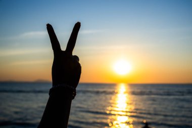 Siluetli el, okyanusun üzerinde parlayan bir günbatımına karşı barış işareti yapıyor. Altın ışık suya yansıyor.