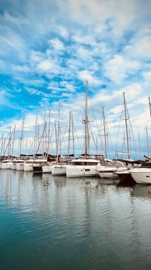Sakin sularda demirlemiş yelkenli ve yatları olan sakin bir marina, dağınık bulutlarla dolu canlı mavi bir gökyüzüne karşı batıyor..