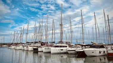 Yelkenli ve yatlarla dolu hareketli bir marina, durgun sulara kenetlenmiş, dağınık bulutlarla dolu mavi bir gökyüzünün altında..