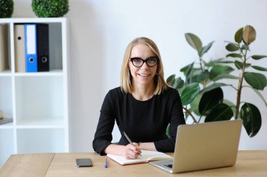 Sırıtkan, gözlüklü, dizüstü bilgisayarına yazı yazan, arka planda bitkiler olan profesyonel bir kadın..